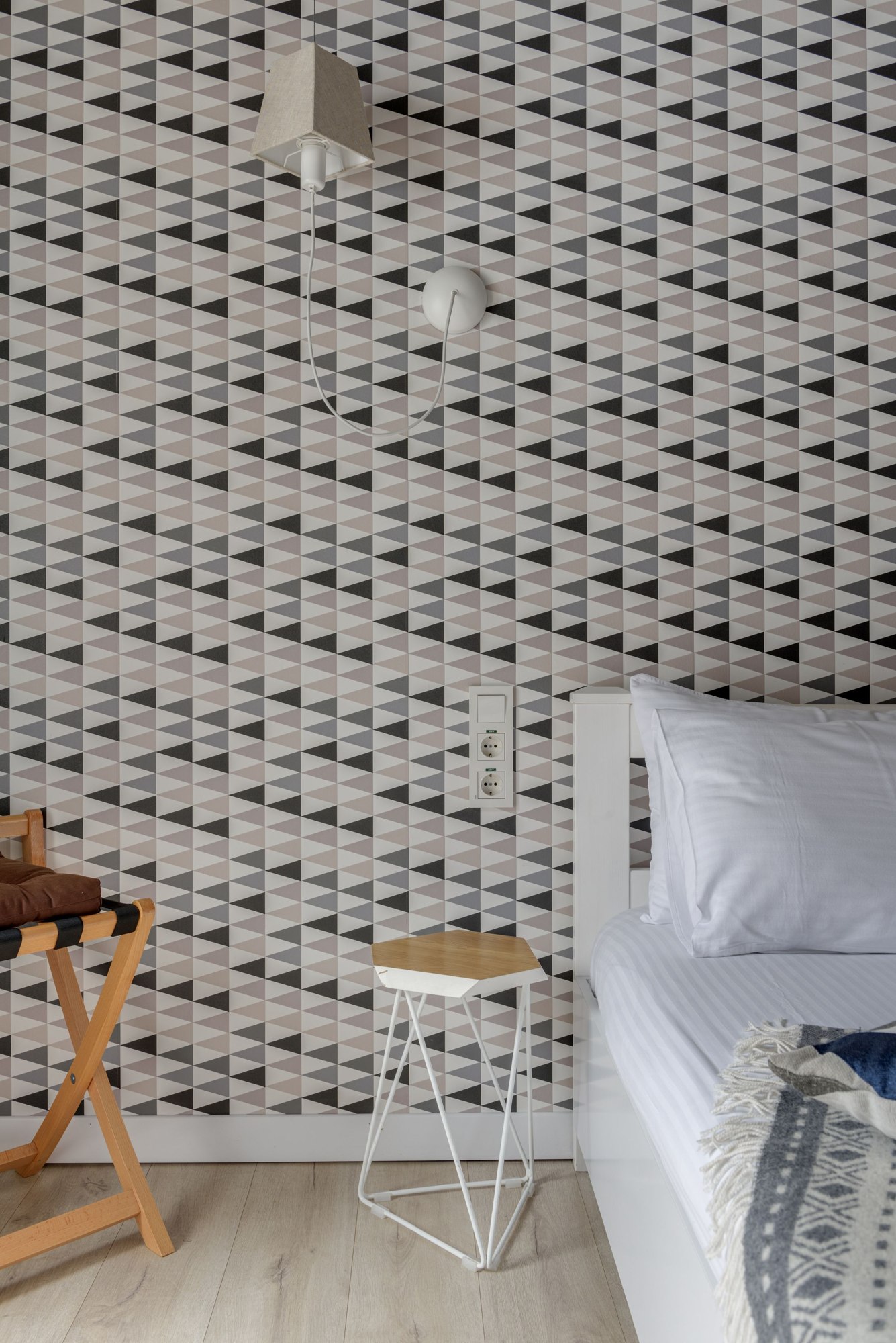 Repeating geometric wallpaper beside a bed and a wooden stool